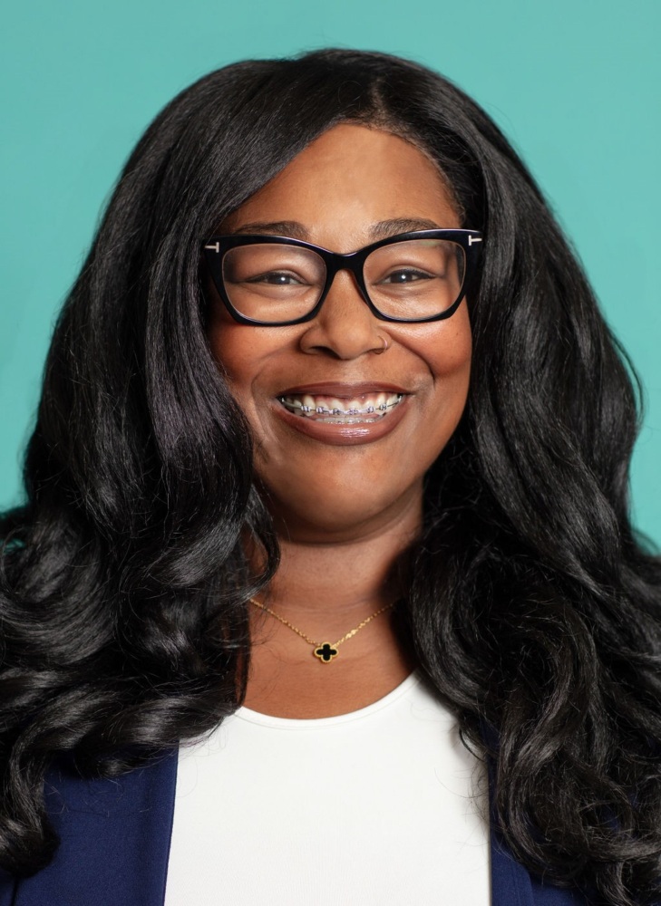 Woman with long black hair, wearing glasses and braces, smiles in front of a teal background. She is dressed in a white top and a dark jacket.